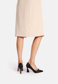 Tacchi alti neri in pelle con punta affusolata e tacco kitten. Presentano un accento decorativo sulla parte anteriore. Indossati con una gonna nuda.