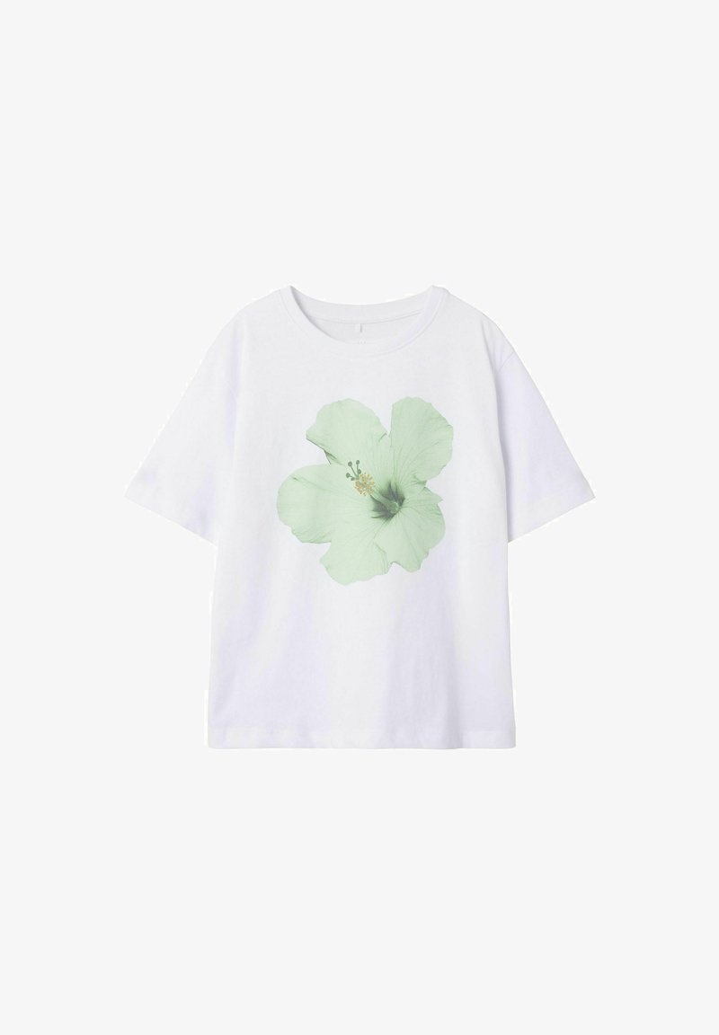 Balta kokvilnas T-kreklā ar gaiši zaļas hibiska zieda grafiku priekšpusē. Ievērojama klasiskā apaļā kakla izgriezuma un īsām piedurknēm.