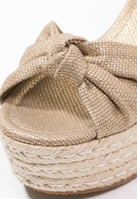 Sandalo in tessuto dorato con tacco a wedge, dettagli con cinturino annodato e suola in juta intrecciata.