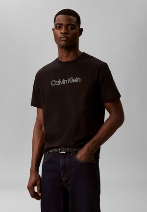 Mężczyzna noszący czarną koszulkę Calvin Klein i ciemne dżinsy, z jedną ręką w kieszeni, stoi na tle jednolitego szarego tła.