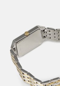 Rektangulär klocka i rostfritt stål med gulddetalj på kronan, armband av silver- och guldmetallkedjor, slät yta och graverad baksida.