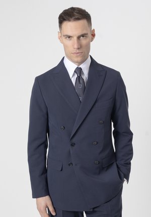 Hombre vistiendo un traje cruzado de color azul marino oscuro con una camisa blanca y una corbata estampada, posando frente a un fondo claro y sencillo.