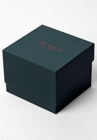 Tumši zaļš dāvanu kastē ar vāku, ar sarkano "TED BAKER LONDON" tekstu augšā, gluda tekstūra un kvadrātveida dizains.