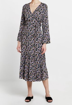 Kobieta ubrana w czarną midi sukienkę w kwiaty z długimi rękawami, dekoltem w serek oraz czarne, otwarte klapki wsuwane.