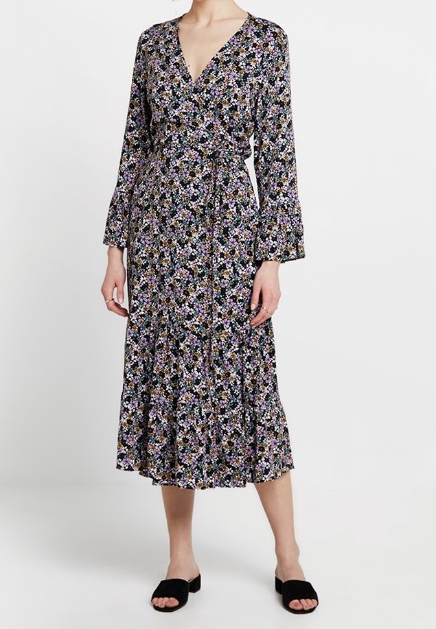Femme portant une robe midi noire à motifs floraux avec manches longues, un décolleté en V et des chaussures noires ouvertes à enfiler.
