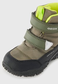 Bota de color verde oliva con dos correas ajustables de Velcro, puntera de goma negra y suela texturizada. Presenta el logo plateado de GEox y un forro de color verde brillante.
