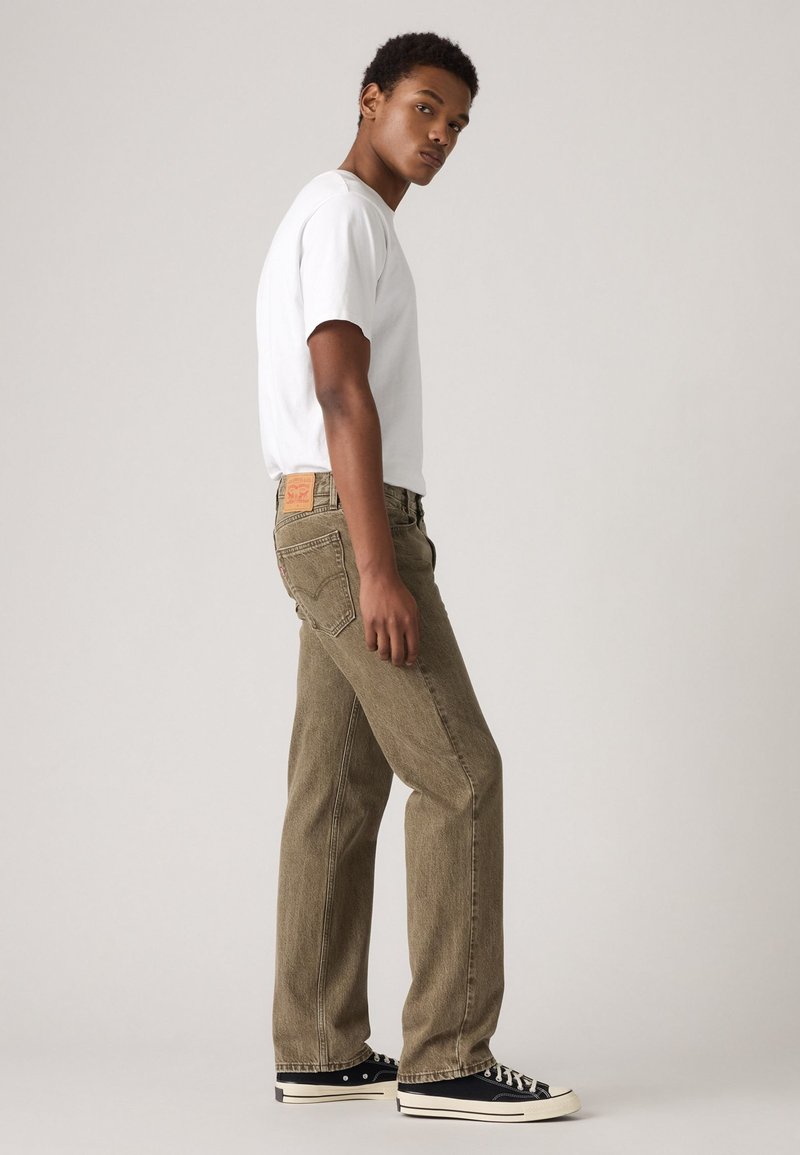 Oliwkowe dżinsy o klasycznym prostym kroju, z skórzaną naszywką marki z tyłu, w połączeniu z białą koszulką i czarnymi trampkami.