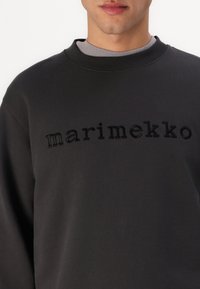 Tmavě šedá mikina s vyvýšeným černým nápisem "marimekko" na hrudi, s kulatým výstřihem a jemným, texturovaným povrchem.