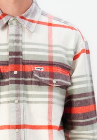 Camisa de franela de cuadros en gris, blanco y naranja. Presenta botones a presión, un bolsillo en el pecho y una etiqueta con el logo de Wrangler en el bolsillo.