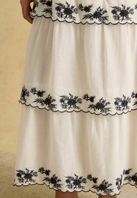 Biela ľanová sukňa s čiernou kvetinovou výšivkou pozdĺž zvlneného okraja, s tromi vrstvenými pásmi zdôrazňujúcimi dizajn.