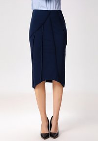 Jupe crayon marine en tissu extensible, pr ésentant un ourlet asymétrique, des coutures diagonales et une texture lisse. portée avec des talons noirs.