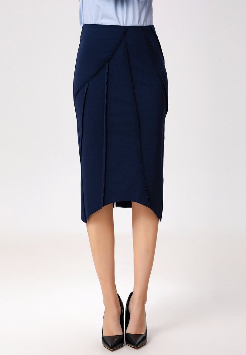Jupe crayon marine en tissu extensible, présentant un ourlet asymétrique, des coutures diagonales et une texture lisse. portée avec des talons noirs.