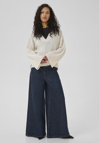 Pull en tricot crème surdimensionné avec un col en V, manches cloche côtelées ; associé à un jean large bleu foncé ; texture lisse, coupe décontractée.