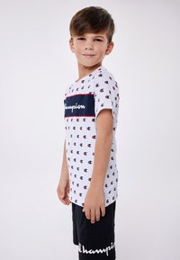 White short-sleeve t-shirt with red, blue, and black Champion logo pattern. Navy horizontal stripe with "Champion" logo across the chest.