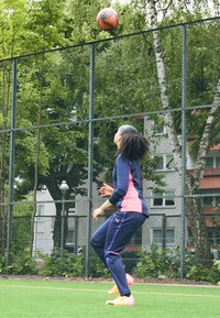 Osoba v námorníckom a ružovom športovom oblečení stojí na trávnatej ploche a pozerá sa hore na oranžovú a čiernu futbalovú loptu nad plotom.