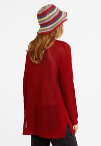 Femme aux longs cheveux roux portant un pull en tricot rouge et un chapeau seau en crochet rayé multicolore, vue de dos.