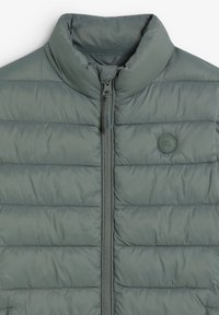 Chaqueta acolchada gris con un cuello alto, acolchado horizontal y cremallera en el frente. Presenta un pequeño logo en el pecho y una textura suave.