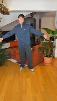 Blue zip-up sweatshirt, black pants, and silver shoes; standing in a living room with a brown couch, plants, and a Christmas tree.