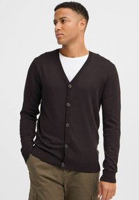 Cardigan marron en tissu tricoté, doté d'un col en V, d'une fermeture à boutons et de poignets et d'ourlet côtelés. Porté par-dessus une chemise blanche.