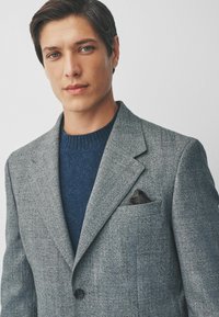 Grau strukturierter Blazer, der über einem marineblauen Sweater getragen wird, mit einem Einstecktuch in dunklen Tönen. Der Blazer hat Revers und einen einzelnen Knopf.