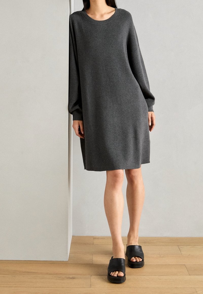 Robe en maille grise à manches longues et col rond, portée avec des sandales compensées noires, sur un sol en bois devant un fond clair.