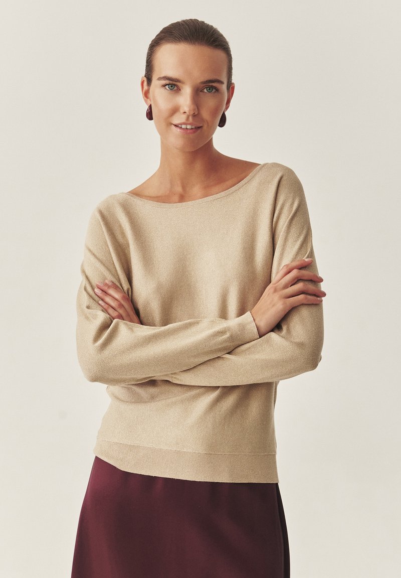 Bež pulover z dolgimi rokavi in širokim izrezom, z rebrastimi manšetami in mehko teksturo, kombiniran z bordo krilom.