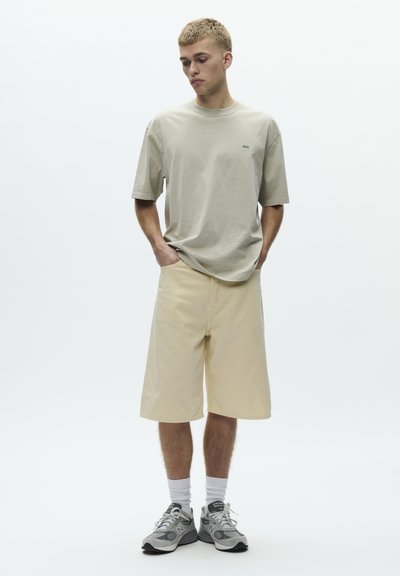 Młody mężczyzna stoi z rękami w kieszeniach, ubrany w beżową, oversize'ową koszulkę, beżowe szorty sięgające do kolan, białe skarpetki i szare sneakersy.
