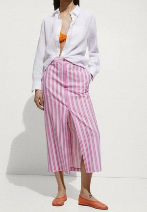 Femme portant une chemise blanche boutonnée, un haut orange, une jupe midi rayée rose et blanche avec une fente frontale, et des ballerines orange, debout devant un fond blanc.