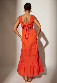 Femme aux cheveux relevés portant une robe maxi orange sans manches avec des volants, un nœud dans le dos et un détail découpé floral, debout les mains sur les hanches.