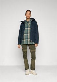 Veste imperméable marine, chemise à carreaux verts, pantalon olive avec des accents en panneau, et chaussures beiges. Vêtements d'extérieur au design fonctionnel.