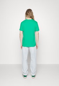 Vihreä puuvillapaita lyhyillä hihoilla ja pyöreällä kaula-aukolla, yhdistettynä vaaleansinisiin farkkuihin ja vihreillä yksityiskohdilla varustettuihin kenkiin, nähtynä takaa.