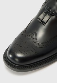 Black leather shoe with a polished finish, featuring brogue detailing, a decorative bow, and a ribbed sole for added texture.