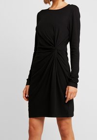 Svart långärmad klänning i mjukt tyg, med en snodd knutdetalj vid midjan. Designen är figurnära, med rundad halsringning och slät textur.