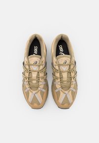 Ett par beige och tan ASICS-sneakers sedda ovanifrån på vit bakgrund, som visar snörning och varumärkeslogotyp inuti.