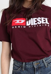 Burgunderfärgad bomull t-shirt med en stor vit och röd "DIESEL"-logotyp och texten "DENIM DIVISION", kombinerad med blå denimshorts.