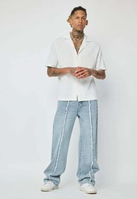 Chemise blanche à manches courtes et boutonnage, avec un motif texturé, associée à un jean large bleu clair avec des coutures latérales blanches contrastantes.