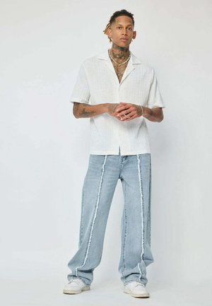Chemise blanche à manches courtes et boutonnage, avec un motif texturé, associée à un jean large bleu clair avec des coutures latérales blanches contrastantes.