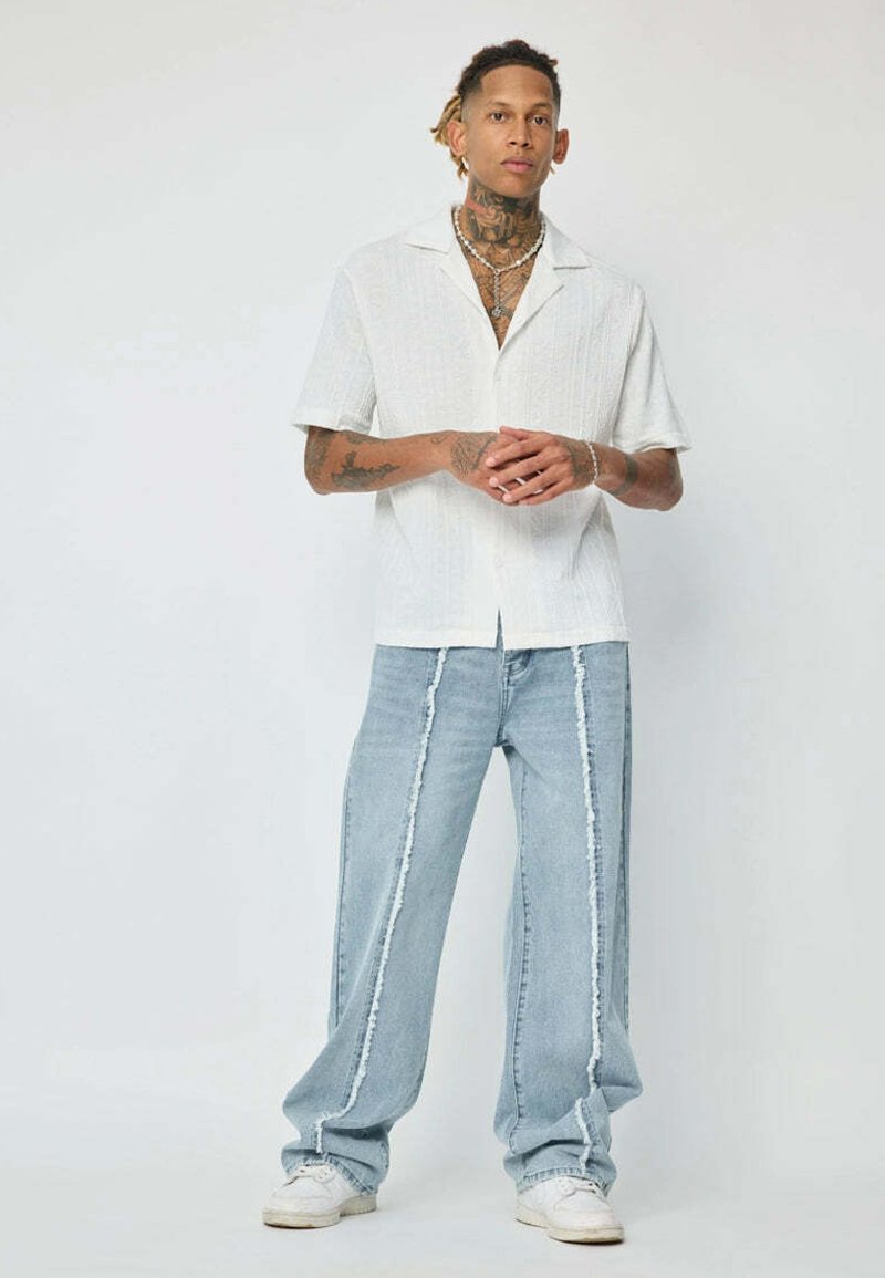 Chemise blanche à manches courtes et boutonnage, avec un motif texturé, associée à un jean large bleu clair avec des coutures latérales blanches contrastantes.