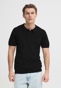 Polo noir à manches courtes, avec un col classique et une patte de boutonnage à trois boutons. Porté avec un jean bleu clair.