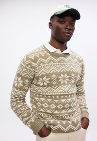 Maglione beige a maglia con motivi geometrici bianchi, dotato di scollatura rotonda e polsini a coste. Il modello indossa una camicia bianca con colletto sotto.