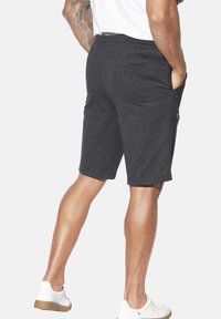 Mann in dunkelgrauen Shorts mit Händen in den Taschen, weißen Turnschuhen und einem weißen Hemd, der vor einem schlichten hellen Hintergrund steht.
