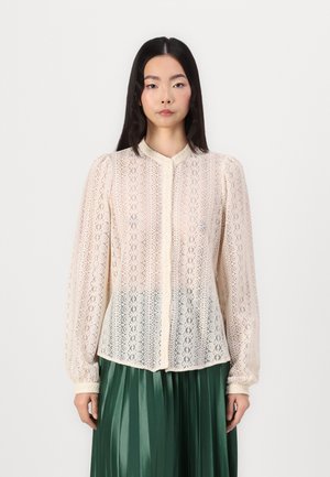 Lace button-up blouse in cream with a floral pattern, long sleeves, mandarin collar, and sheer texture, paired with a pleated green skirt.