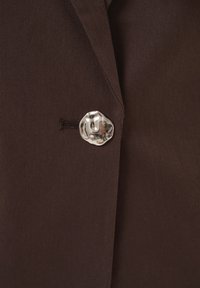 Tissu de blazer marron avec un seul bouton métallique argenté brillant de forme irrégulière, fixé à travers une boutonnière cousue.