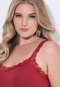 Blonďatá žena s vlnitými vlasy má na sobě červenou camisolku s krajkovým lemováním a geometrické náušnice v růžové a zlaté barvě, dívá se přímo do kamery.