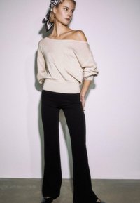 Ženska stoji z rokami za hrbtom, oblečena v bež pulover brez ramo, črne hlače, črne čevlje in črno-belo rutko s potiskom.