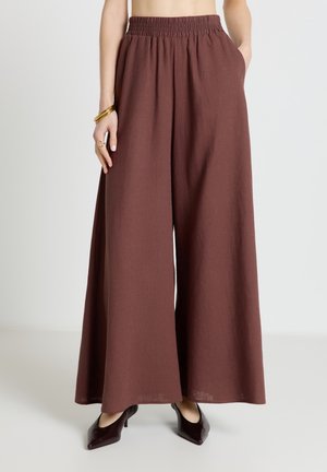 Donna che indossa pantaloni marroni a vita alta e gamba larga con vita elasticizzata, abbinati a décolleté scuri con punta appuntita e braccialetti dorati al polso sinistro.