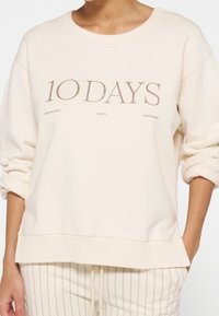 Sweat-shirt beige clair avec une coupe décontractée, des poignets et un ourlet côtelés. Texte graphique frontal en marron : « 10 DAYS UNEXPECTEDLY SIMPLE AMSTERDAM ».
