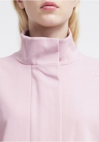 Pink high-neck garment with a smooth texture and minimal design. Features a folded collar and stitching detail, creating a sleek silhouette.