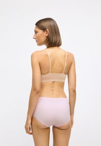 Vrouw met kort bruin haar, gekleed in een beige bh met verstelbare schouderbandjes en lichtroze high-waisted ondergoed met kantafwerking, staand tegen een witte achtergrond.