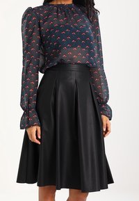 Jupe en cuir noir à plis associée à un chemisier transparent avec un motif floral en bleu foncé et rouge, manches longues bouffantes.